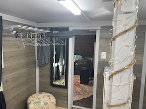 A room with a wooden floor, a hanging clothes rack, a mirror, a small stool, and a doorway leading to another room. - Charming Rustic Tiny Home – Private 1 Ac Oasis - Yulee, FL (Yulee)