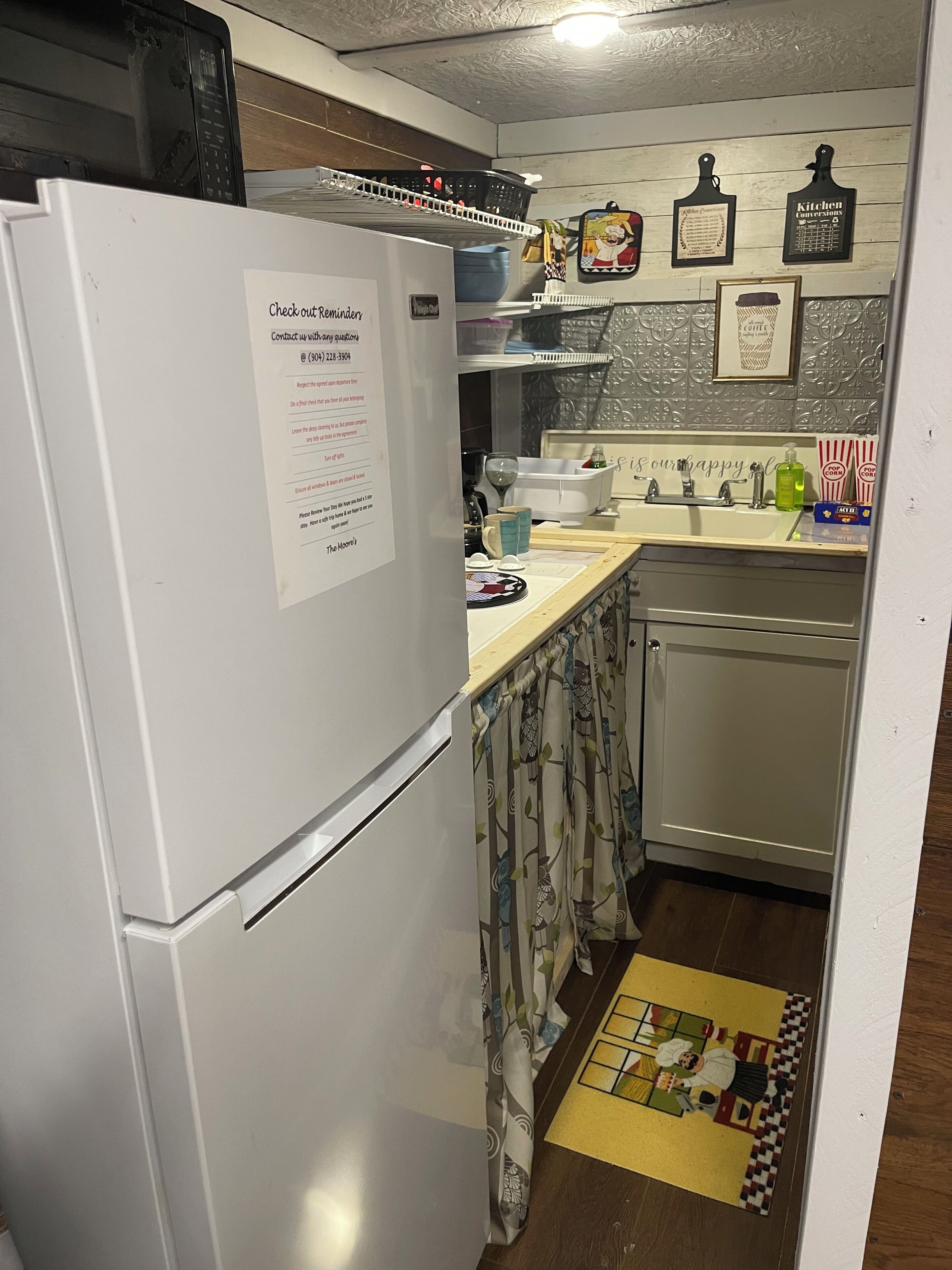 A kitchen with a white refrigerator, a sink, and a coffee machine. There is a note on the refrigerator, a patterned rug, and kitchen decorations on the wall.