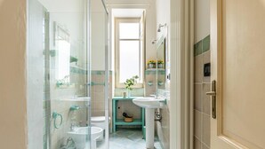 A bathroom with a glass shower enclosure, a toilet, a sink, and a small shelf with toiletries. - Zisa Gardens of Palermo (PALERMO)