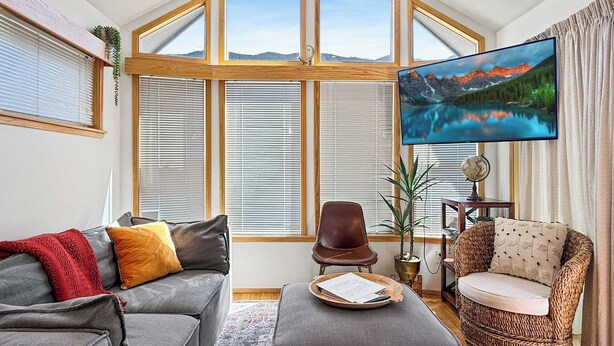 A living room with a grey sofa, a brown leather chair, a wicker armchair, a flat-screen TV, and a large window with blinds. - Lake Sutherland Tiny Home, Heaven (Port Angeles)