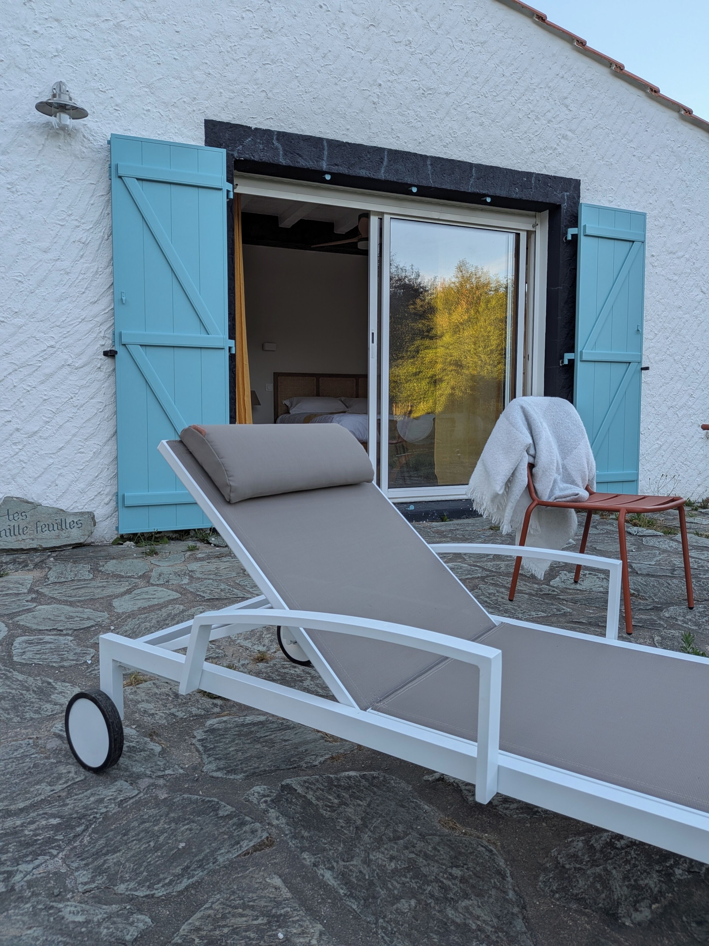 Une chambre avec un lit, une fenêtre avec des volets bleus et un fauteuil.