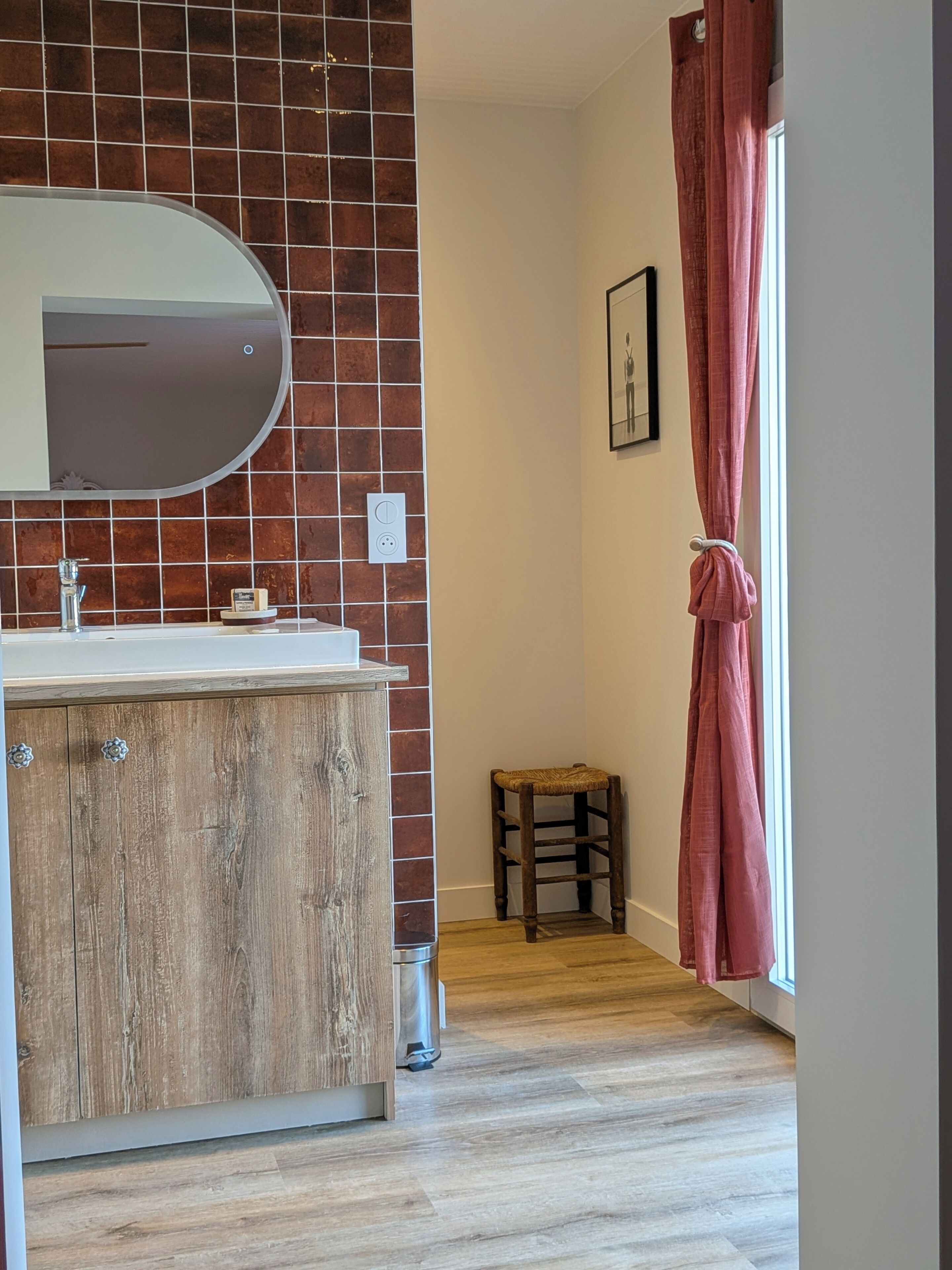 Une salle de bain avec un meuble-lavabo en bois, un miroir rond, un mur carrelé, un petit tabouret et une fenêtre avec un rideau.