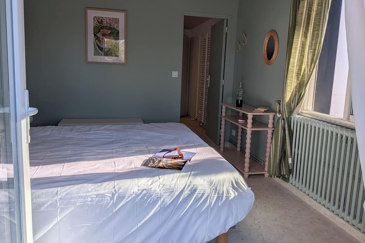 A bedroom with a bed, a bedside table with a bottle, a framed picture, and a window with curtains.