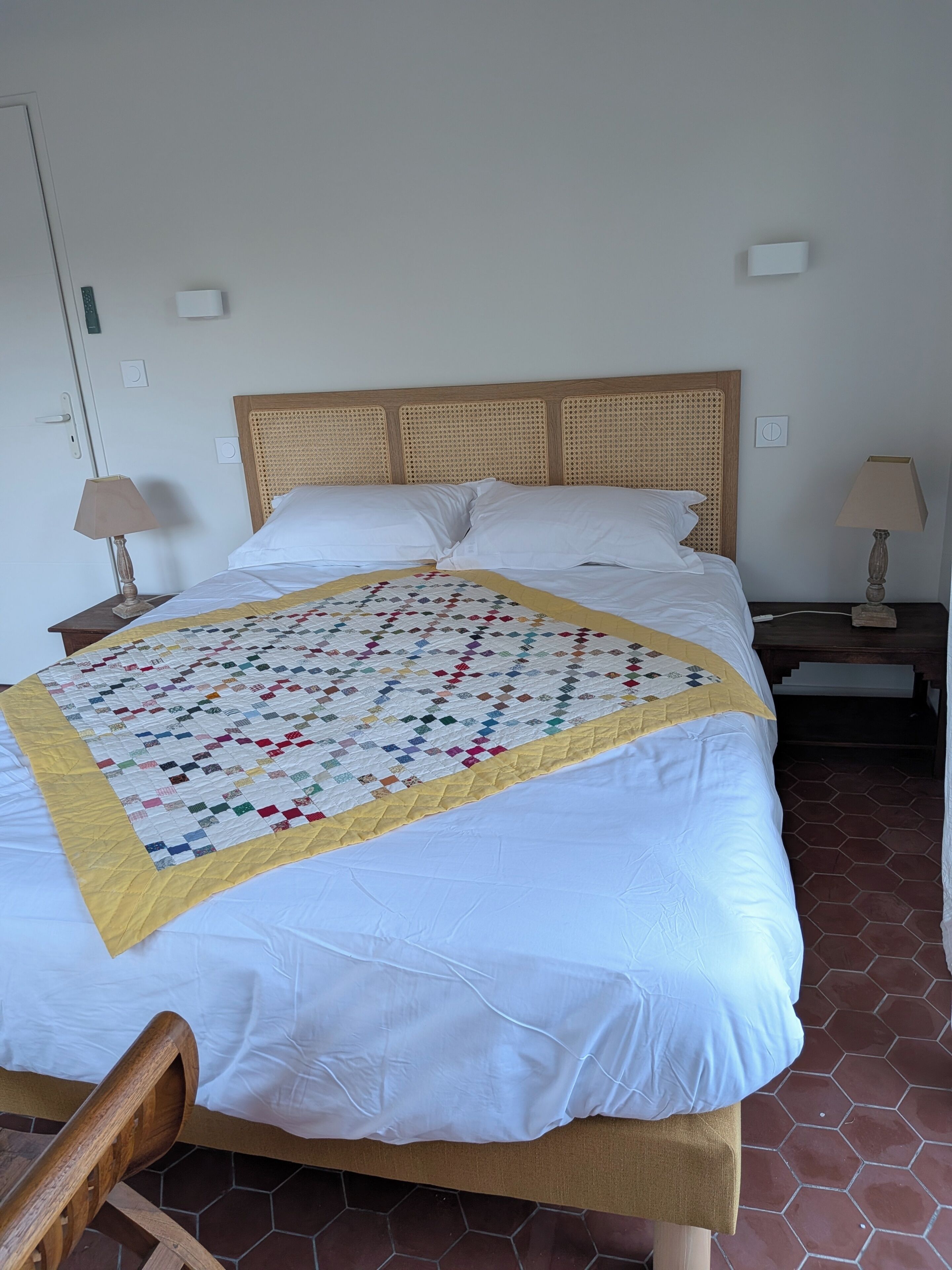 Un letto con una coperta colorata, una testiera in legno, cuscini bianchi, due lampade ai comodini e un pavimento piastrellato.