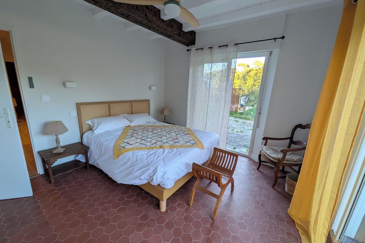 A bedroom with a bed, a chair, a nightstand, a lamp, and a window with curtains.