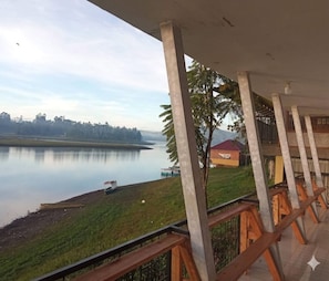 A view from a covered balcony overlooking a calm lake with a distant shoreline, a small boat, and a building with a red roof.