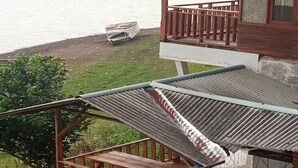 Una terraza de madera con techo metálico, con vistas a un espacio verde con una pequeña mesa y un edificio con balcón.