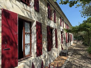 Ein Steinhaus mit roten Fensterläden, einer kleinen Außen-Sitzgelegenheit und einem Kiesweg.