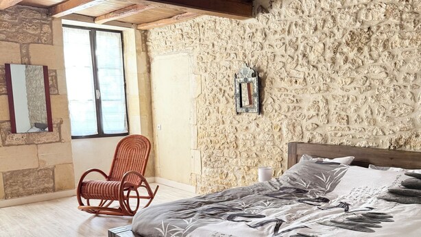 Ein Schlafzimmer mit einem hölzernen Bett, einem Korbstuhl, Steinwänden und einem großen Fenster.