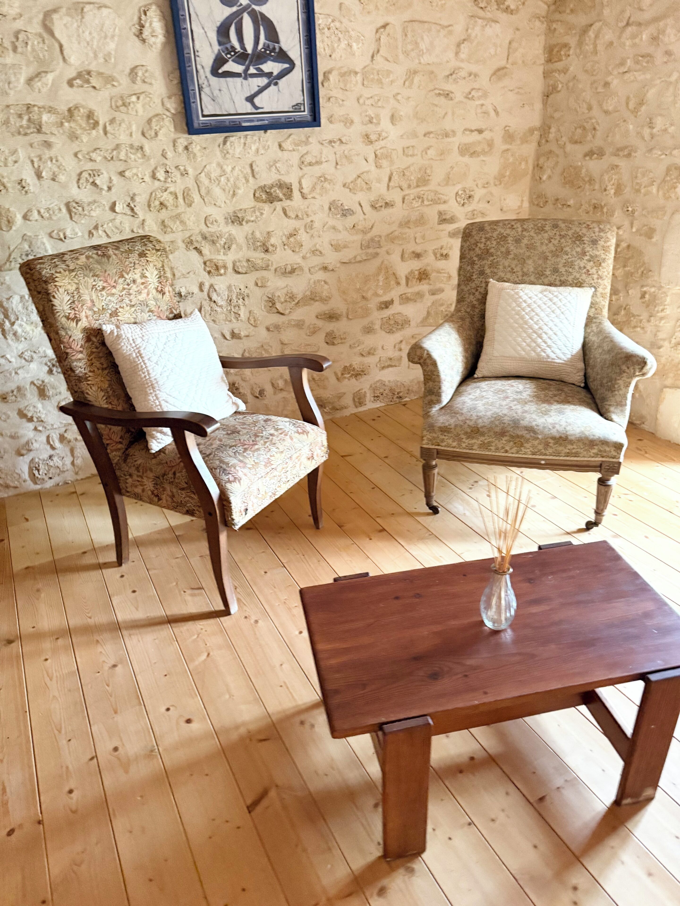 Two armchairs with patterned upholstery and white cushions, a wooden coffee table with a vase, and a stone wall with a framed artwork.
