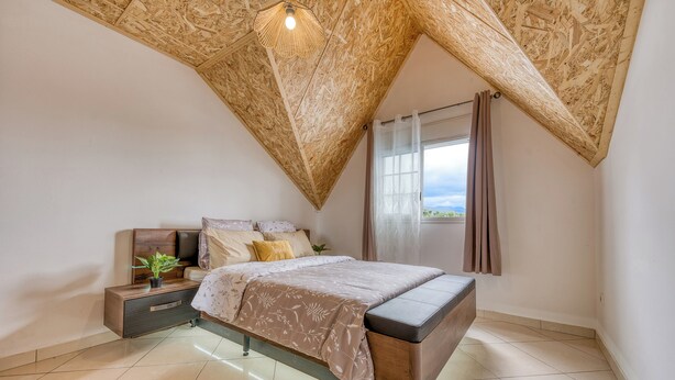 Ein Schlafzimmer mit schrägen Wänden, einem Bett, einem Nachttisch und einem Fenster mit Vorhängen.