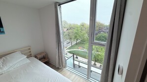 A bedroom with a large window, a bed, a nightstand, and a view of a green park and buildings.