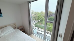 A bedroom with a large window, a bed, a nightstand, and a view of a green park and buildings.
