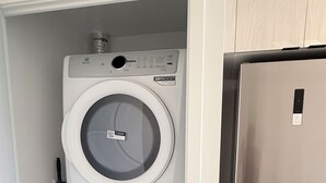 A compact laundry room with a top-loading washer and a front-loading dryer, a broom, and a stainless steel refrigerator.