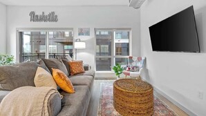 Ett modernt vardagsrum med en grå soffa, en platt-TV, en ottoman i rotting och ett stort fönster.