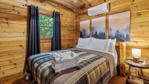 Une chambre dans une cabane en bois, avec un lit, des rideaux, un ventilateur de plafond, un climatiseur et un tableau représentant des arbres.