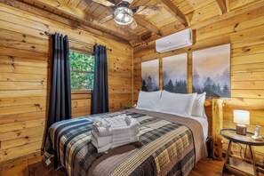 A wooden cabin bedroom with a bed, curtains, a ceiling fan, an air conditioner, and a painting of trees. - Creekfront Escape •Firepit •Hiking • Pet Friendly (Blue Ridge)