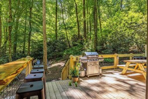 Een terras buiten met een barbecue, zitplaatsen en uitzicht op een bos.