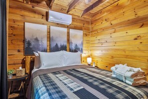Een houten kamer met een bed, nachtkastje, lamp en een raam met uitzicht op bomen.