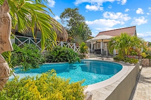 A swimming pool with a stone border, surrounded by tropical plants and a building with a tiled roof.