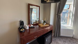 Quarto de hotel com cômoda de madeira, espelho, máquina de café, bandeja com copos, garrafa de champanhe e vista da cidade pela janela.