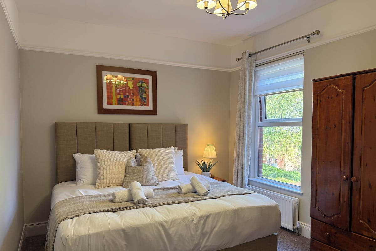 A neatly made bed with pillows and a blanket, a bedside lamp, a window with curtains, and a wooden wardrobe.
