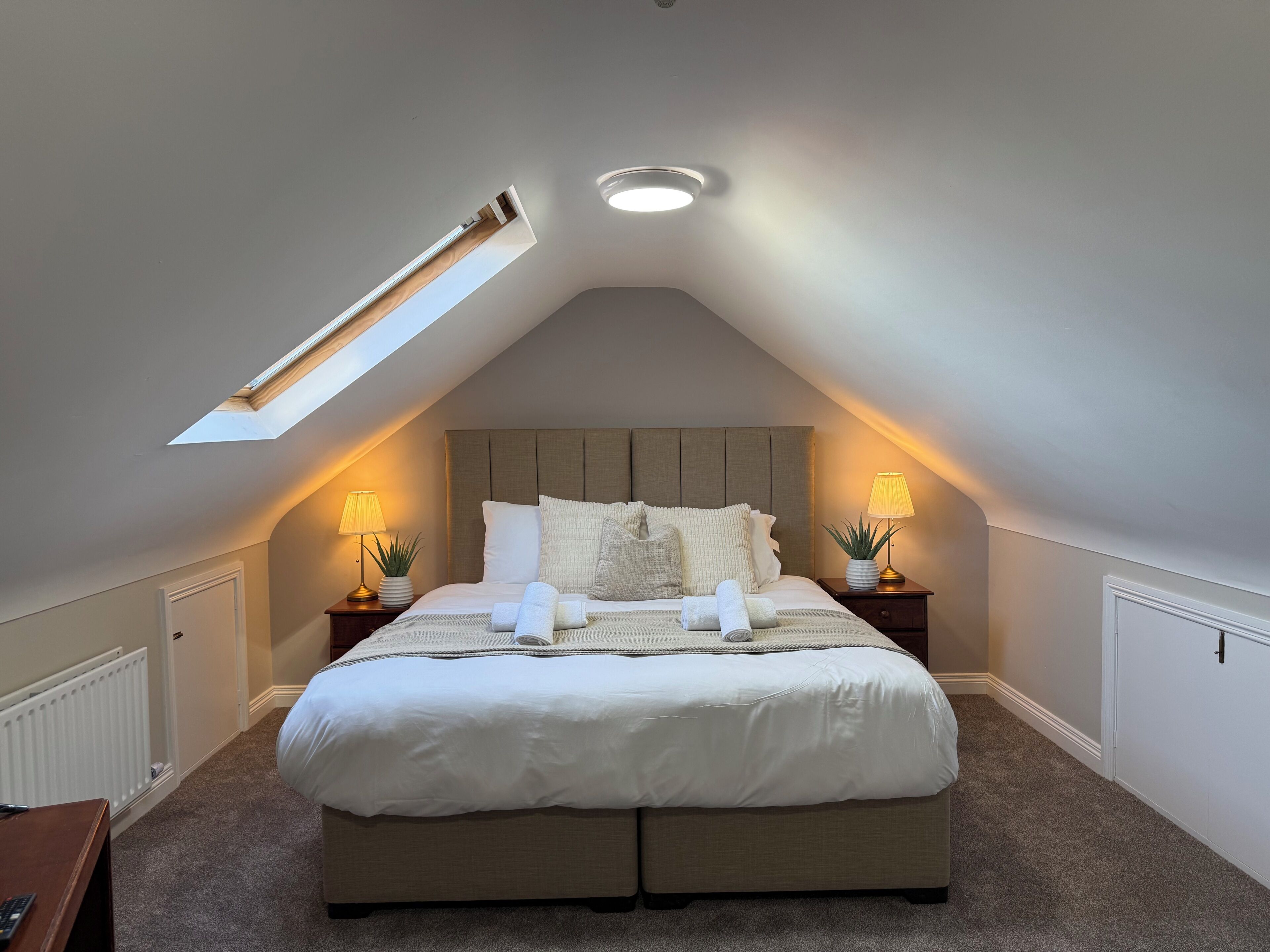 A bedroom with a sloped ceiling, a skylight, a bed with white bedding, two bedside tables with lamps, and a radiator.
