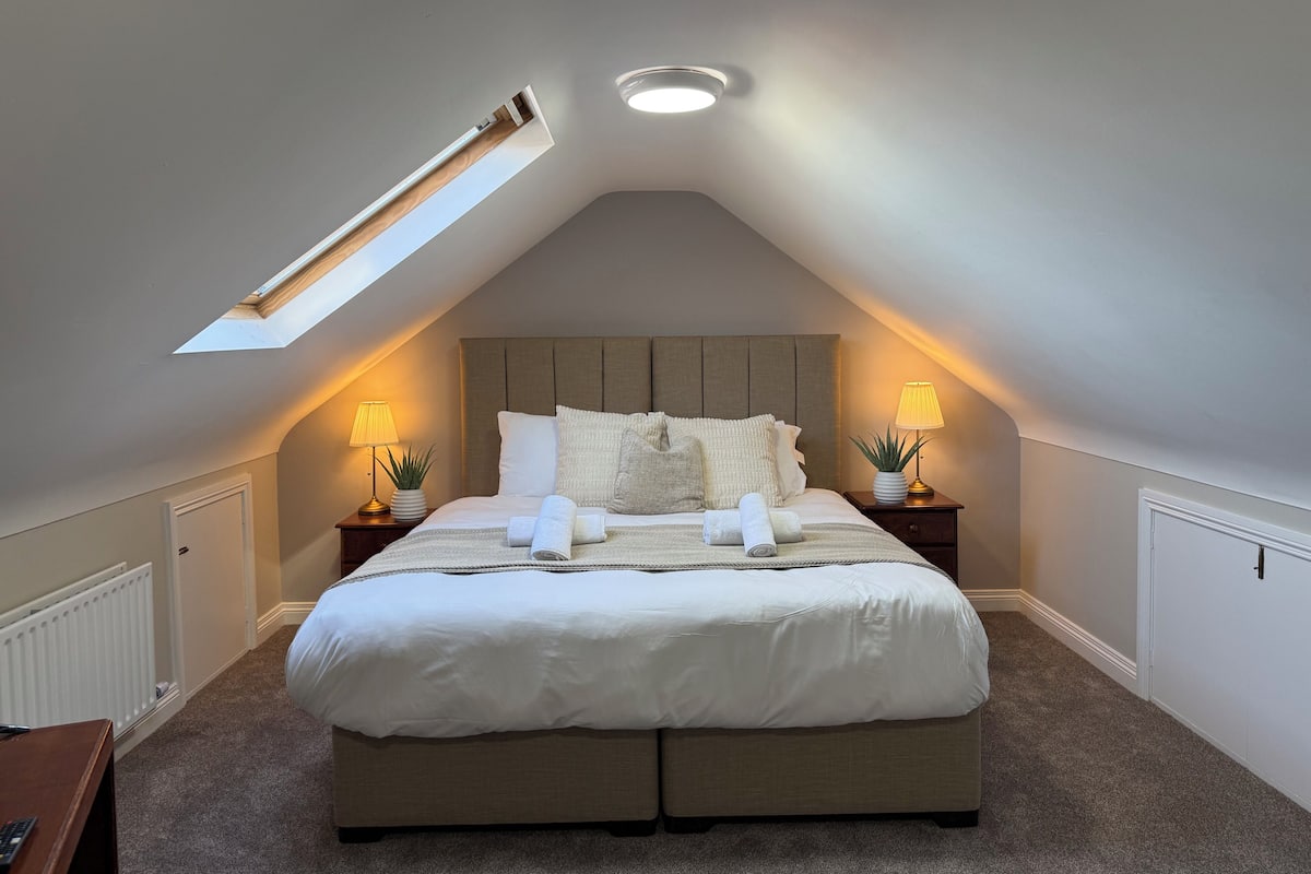 A bedroom with a sloped ceiling, a skylight, a bed with white bedding, two bedside tables with lamps, and a radiator.