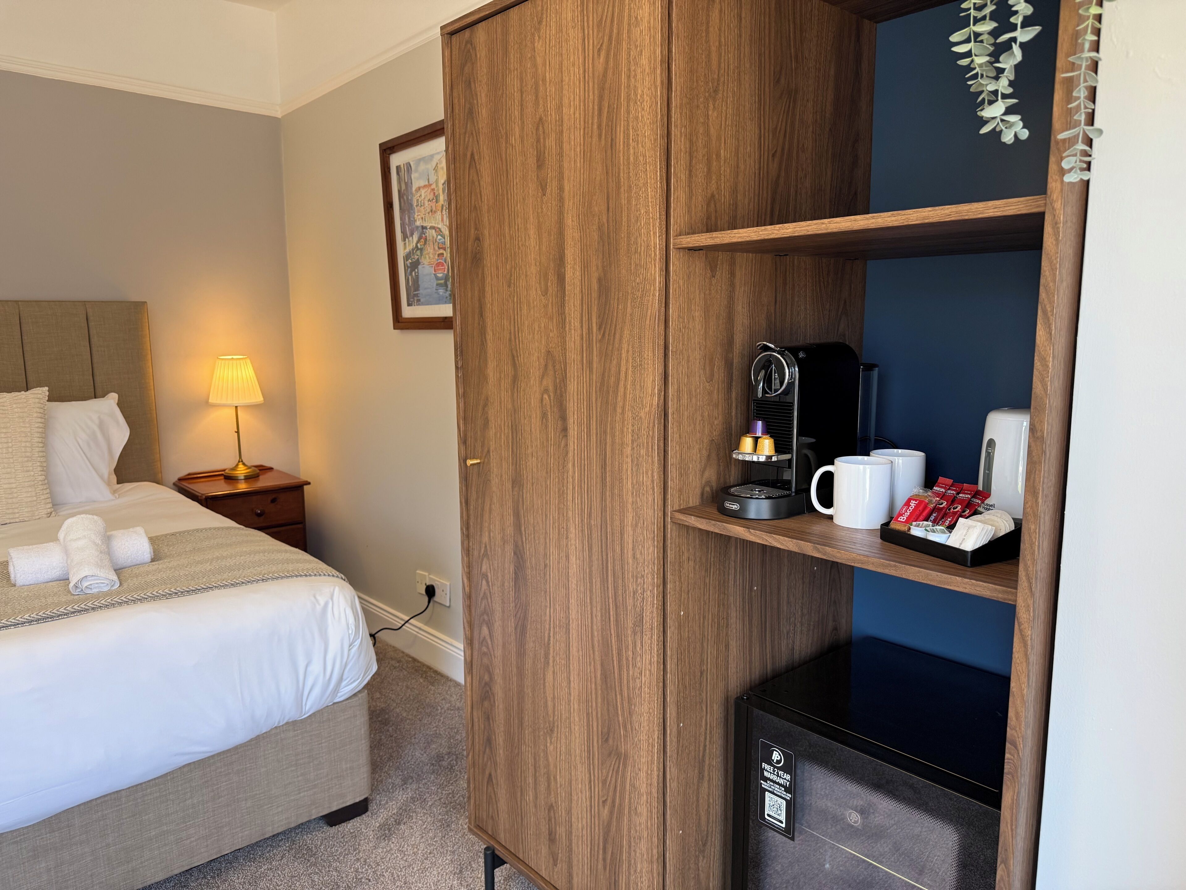 A hotel room with a bed, a nightstand with a lamp, a wooden wardrobe, a coffee machine, and a shelf with coffee cups and packets.