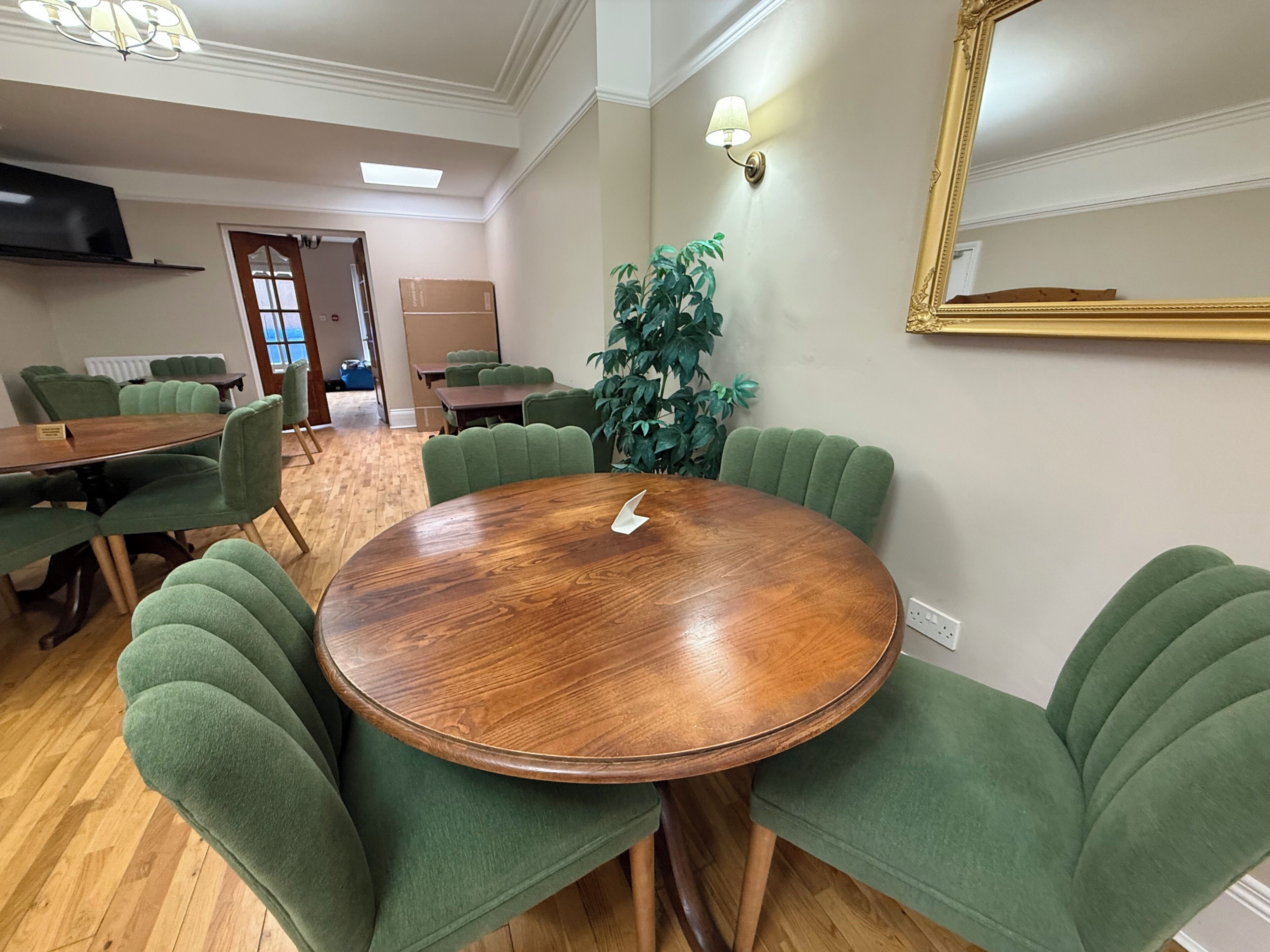 A conference room with a round wooden table, green upholstered chairs, a potted plant, and a wall-mounted mirror.