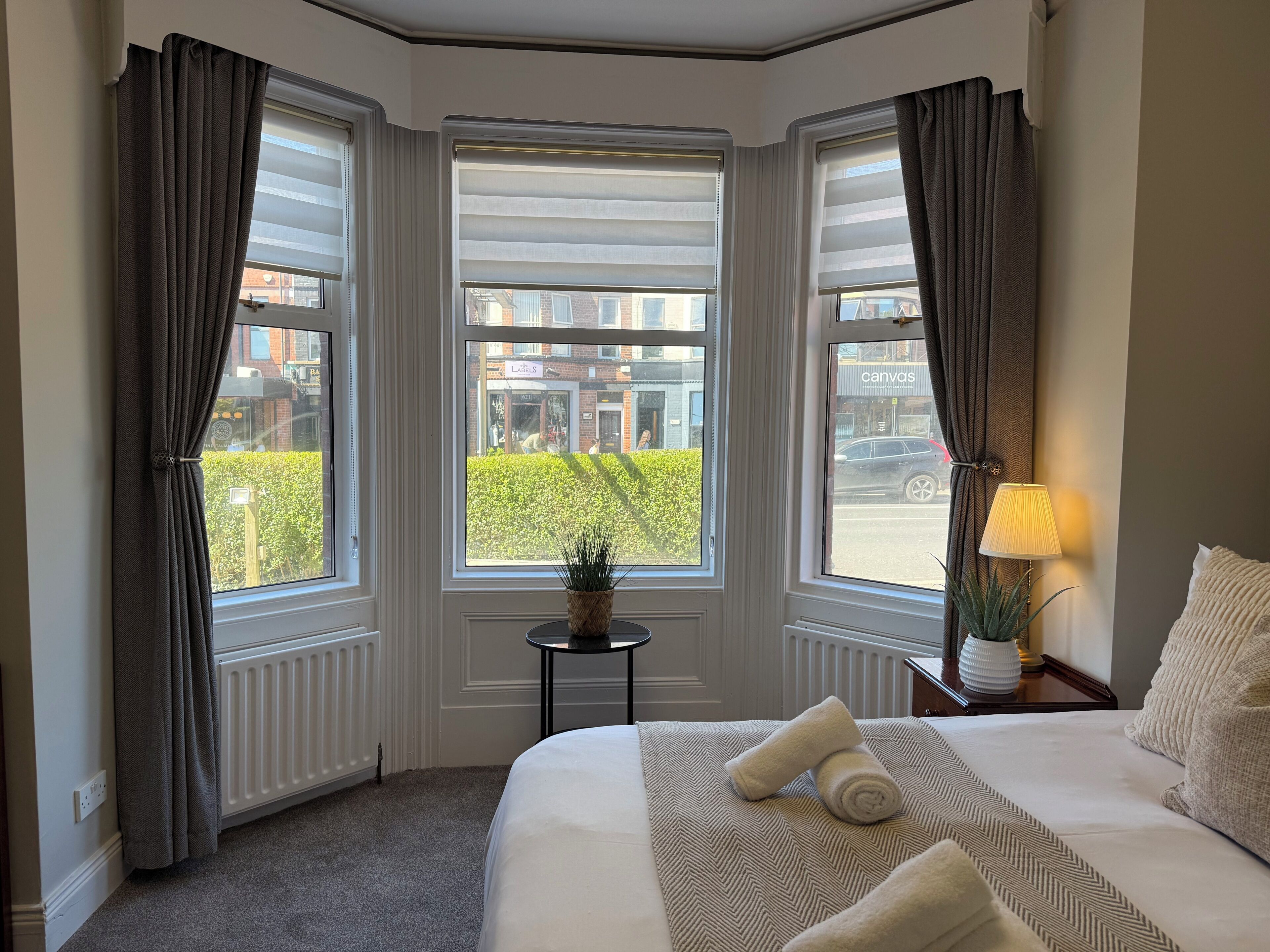 A hotel room with a large bed, a small table with a lamp, and a view of the street through three windows.