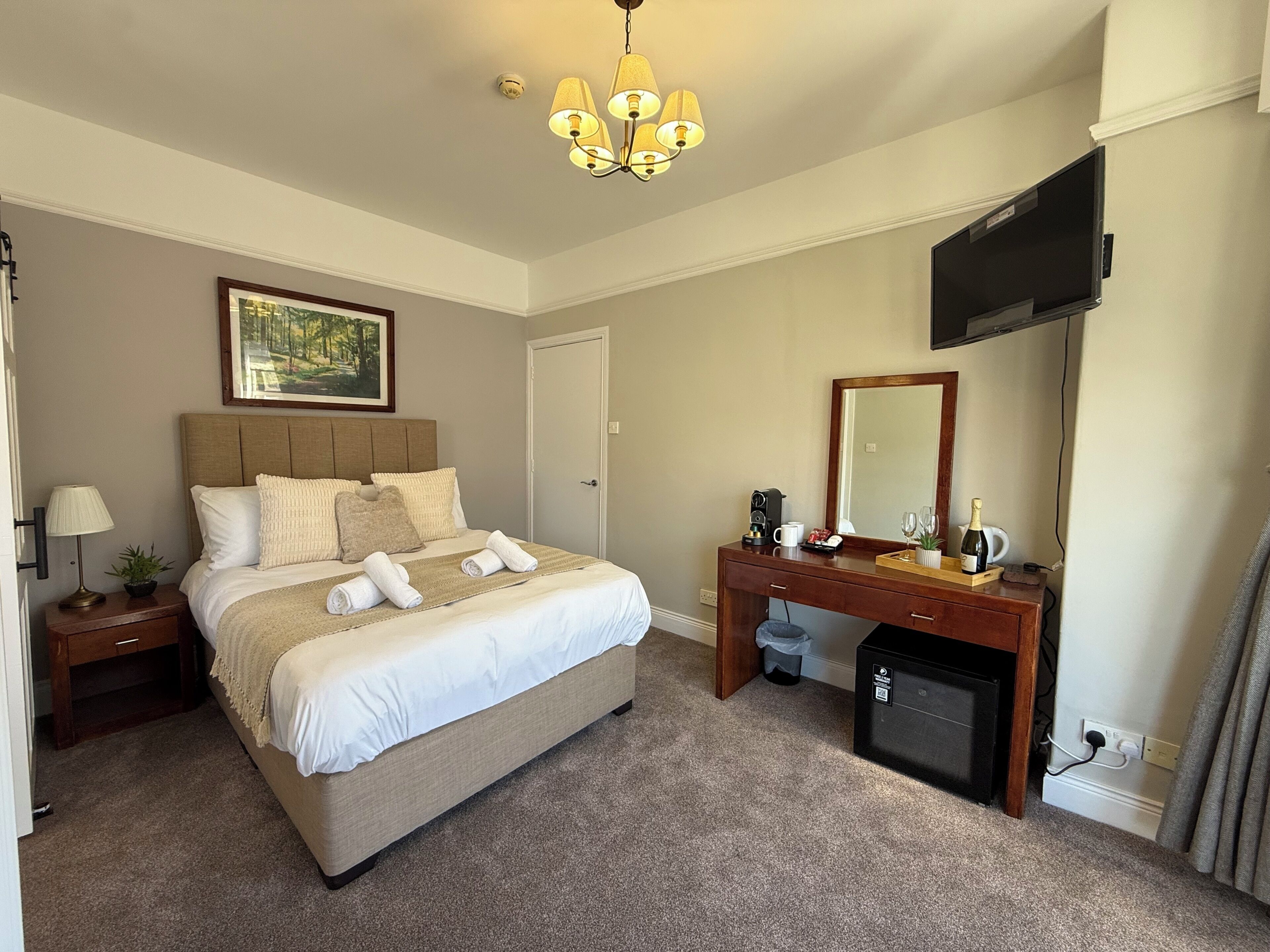 Quarto de hotel com cama, mesa de cabeceira, luminária e televisão na parede.