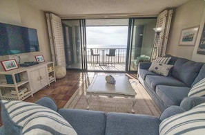 Ein Wohnzimmer mit einer Couch, einem Couchtisch, einem Fernseher, einem Schrank und einem Balkon mit Meerblick.