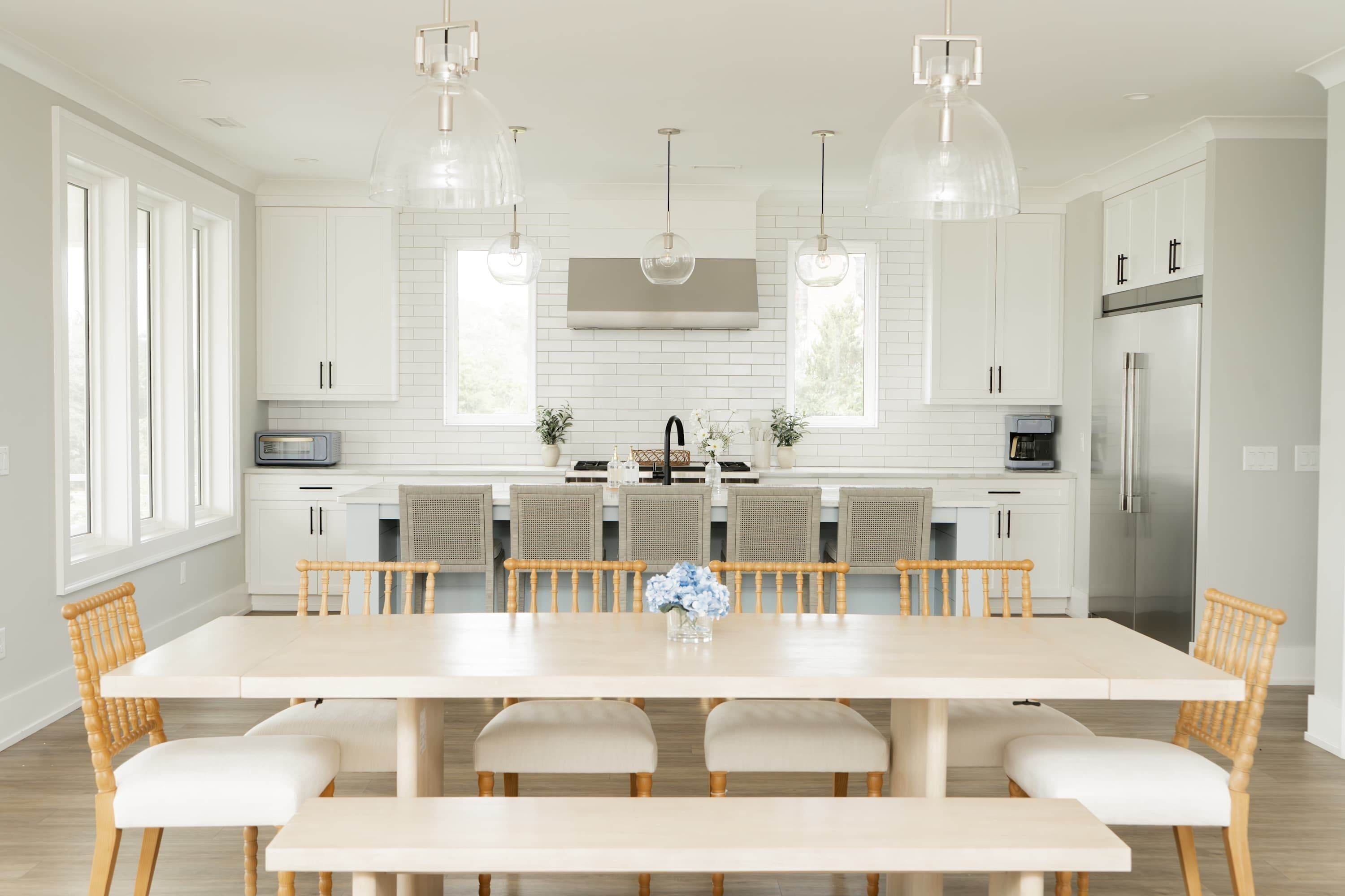 Une cuisine moderne avec des meubles blancs, un îlot central, des luminaires suspendus et un coin repas comprenant une longue table et des chaises.
