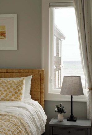A bedroom with a bed, a nightstand with a lamp, a framed picture, and a window with curtains. - 13 Raleigh: Pool, Ocean Views, 2 Houses to Beach (Wrightsville Beach)
