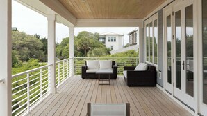 A covered outdoor deck with a table and chairs, a sofa, and a railing overlooking a lush landscape. - 13 Raleigh- Ocean views, Private Pool and more (Wrightsville Beach)