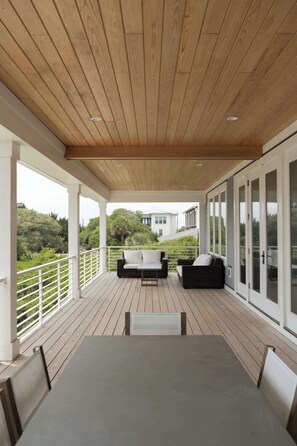 Eine überdachte Terrasse im Freien mit Tisch und Stühlen, einer Sitzecke und einem Geländer mit Blick auf eine üppige Landschaft.