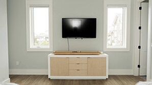 A room with a TV mounted on the wall, a wooden cabinet below, and two windows. - 13 Raleigh- Ocean views, Private Pool and more (Wrightsville Beach)
