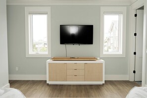 Ein Zimmer mit einem an der Wand befestigten Fernseher, einem Holzkasten darunter und zwei Fenstern.