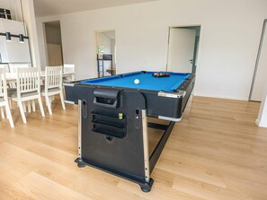 Ein Billardtisch mit schwarzem Tuch, umgeben von weißen Stühlen, in einem Raum mit Holzboden und weißen Wänden.