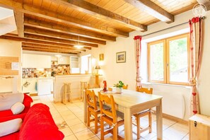 Ein gemütliches Zimmer mit Holzboden, einem Essbereich mit Tisch und Stühlen, einer Küche im Hintergrund und einem Fenster mit Vorhängen.