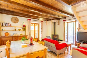Ein gemütliches Zimmer mit Holzbalkendecke, einem Esstisch, Stühlen, einer Couch, einem Fernseher und einem Kamin.