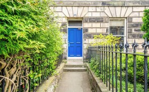 A narrow pathway leading to a blue door, flanked by greenery and a black metal fence. - Beautiful 3 bedroom Georgian flat in Newington area. (Edinburgh)