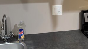 A kitchen with a white countertop, a sink, a faucet, a bottle of Ajax dish soap, and a cabinet with shelves holding white mugs and bowls.