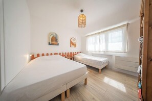 Ein Zimmer mit zwei Betten, einem Vorhangfenster, einer Pendelleuchte und einer Heizkörper.