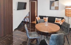 Ein Essbereich mit einem runden Tisch, grau gepolsterten Stühlen, einer Couch mit gemusterten Kissen, einem an der Wand befestigten Fernseher und einer Holztür.