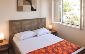 Ein Bett mit Holz Kopfteil, weißer Bettwäsche, einer gemusterten Tagesdecke, einer Nachttischlampe und einem Fenster mit Blick auf Bäume.