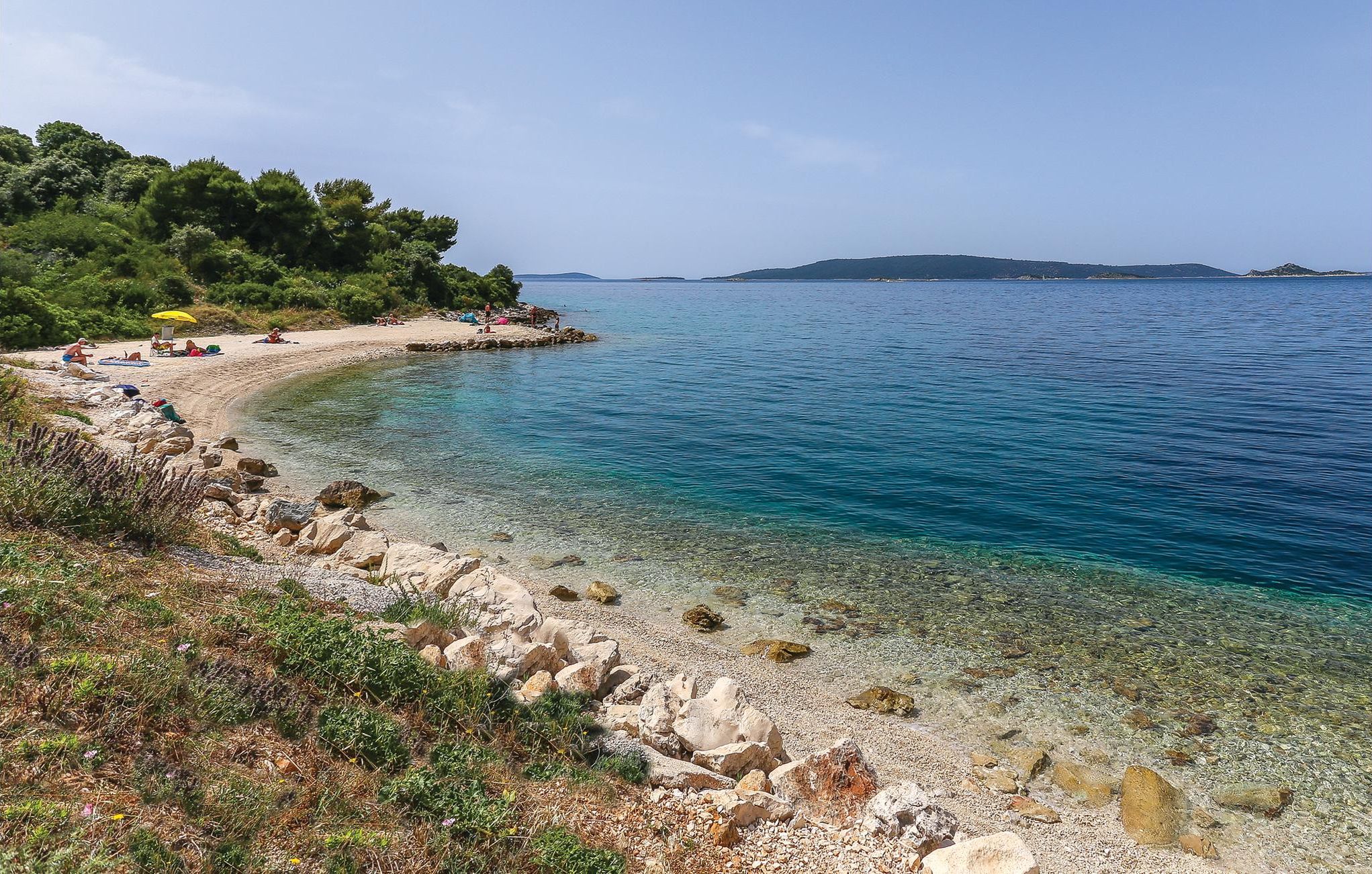 A beach with clear shallow waters, rocky shoreline, and a forested area in the background.