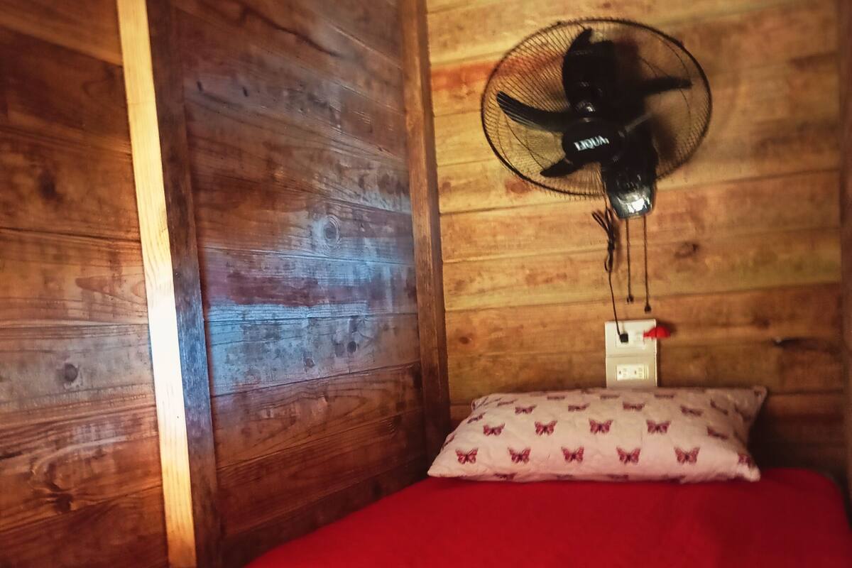 A wooden room with a bed, a wall-mounted fan, and a power outlet.