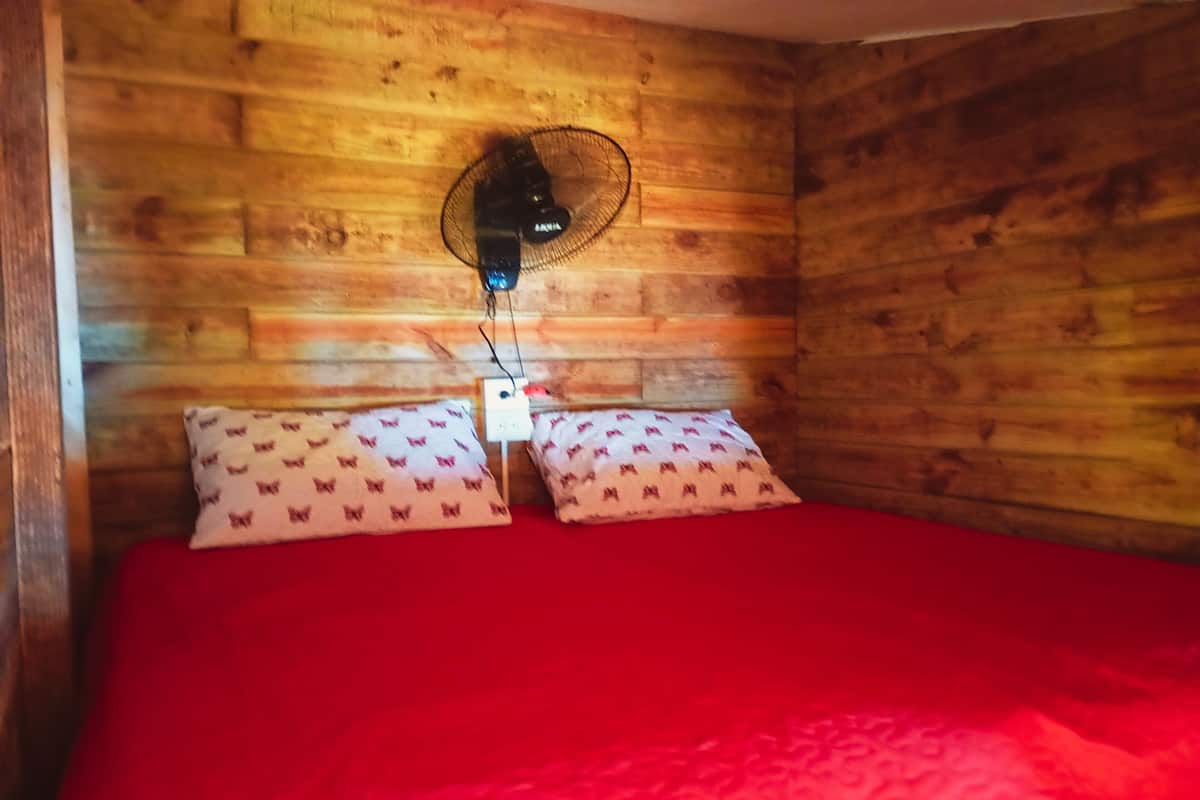 A wooden room with a bed, two pillows, a wall-mounted fan, and a power outlet.